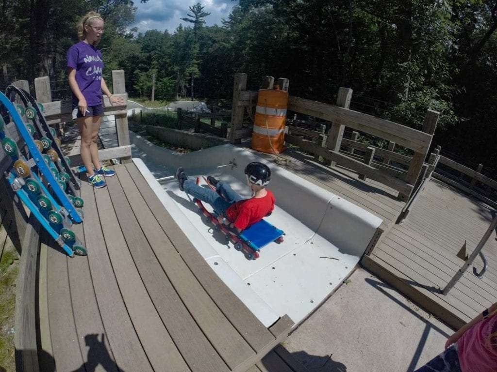 A child in a red helmet enjoys a simulated sledding experience on a dry slide at a park in Muskegon, under the watchful eye of a supervisor.