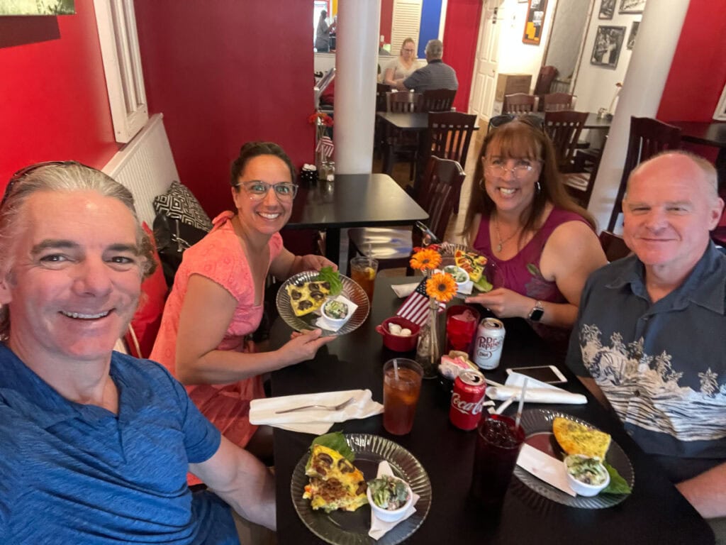 Four smiling adults sit around a table in a lively restaurant, sharing plates of food and drinksโperhaps discussing how to plan a vacation with other families. The bright red walls and cheerful atmosphere add to the fun, with flowers and soda cans on the table.