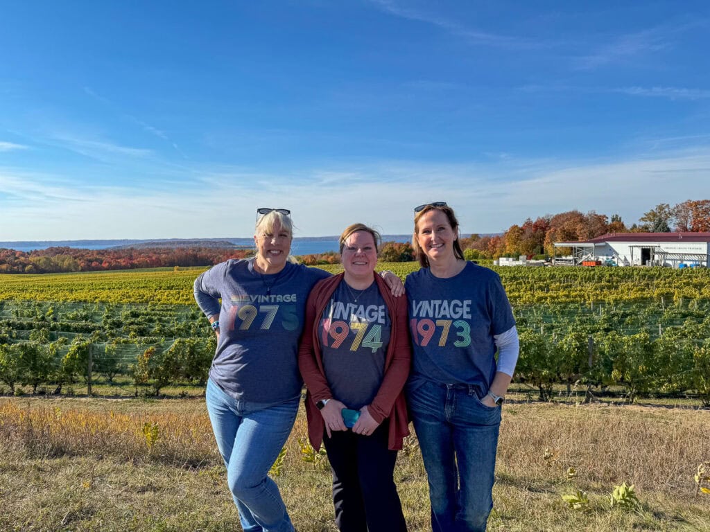 Three women smile in front of a vineyard on a sunny day, wearing matching "VINTAGE 1973" and "VINTAGE 1974" shirts. Autumn foliage and a building hint at the perfect backdrop for multifamily vacation destinations.