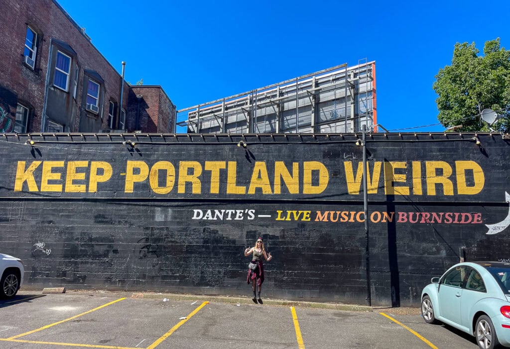 A person stands in a parking lot before a large black mural with yellow text reading "KEEP PORTLAND WEIRD" and smaller text advertising "DANTE'S โ LIVE MUSIC ON BURNSIDE," hinting at Portland's charm as one of many multifamily vacation destinations.