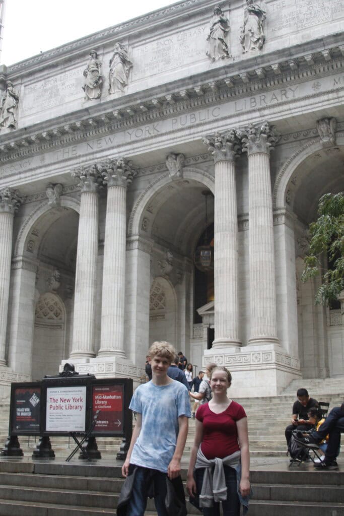 Two people stand in front of the New York Public Library's grand entrance, with its iconic columns and steps. Visitors explore in the background, eager to save money on attractions while absorbing the library's rich history, guided by a sign brimming with fascinating information.