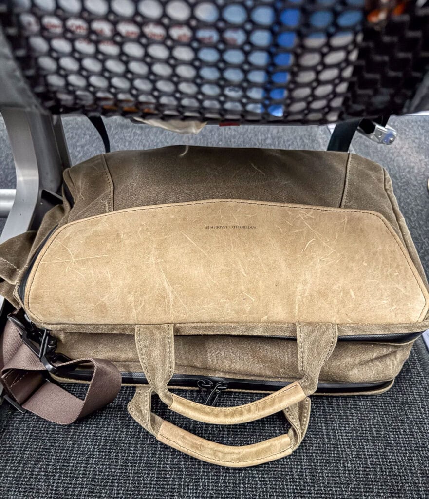 A brown canvas underseat carry-on duffel bag with handles and a shoulder strap rests on a dark carpeted floor beneath an airplane seat, where a mesh pocket and part of the metal frame are visible above.