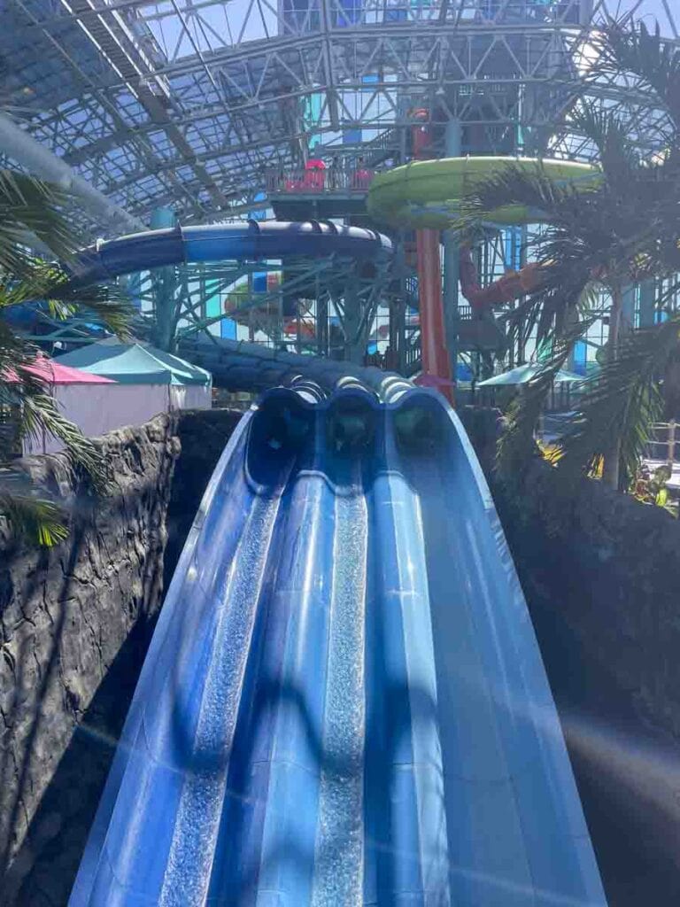 A large indoor water park in Gulf Shores features multiple water slides. Three blue slides run parallel, ending at the bottom. The background showcases green and orange slides, palm trees, and a glass ceiling that allows natural light to illuminate the amusement park.