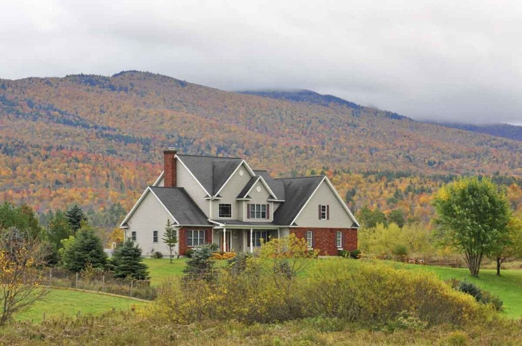 A two-story house with a mix of light gray siding and red brick sits on a lush green lawn, surrounded by trees with autumn foliage. Behind the house, rolling hills covered in colorful fall leaves extend toward the horizon under a cloudy sky—truly one of the best places to visit in the fall.