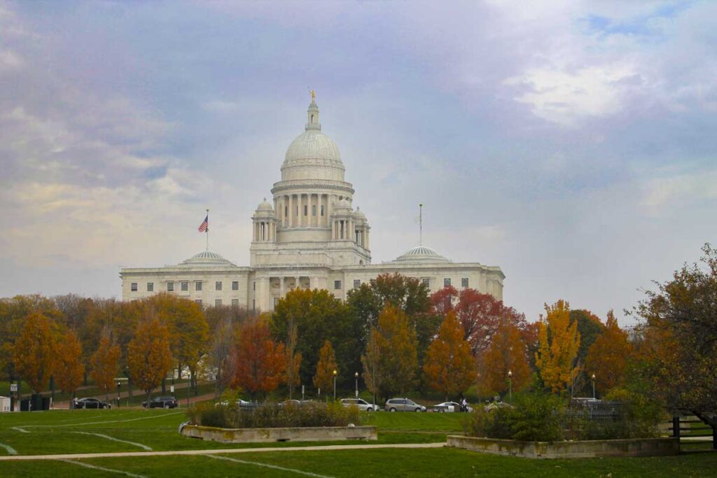 A majestic white-domed government building surrounded by trees in autumn colors stands against a cloudy sky. Cars are parked in front, and well-kept lawns and pathways are visible in the foreground—truly one of the best places to visit in the fall.