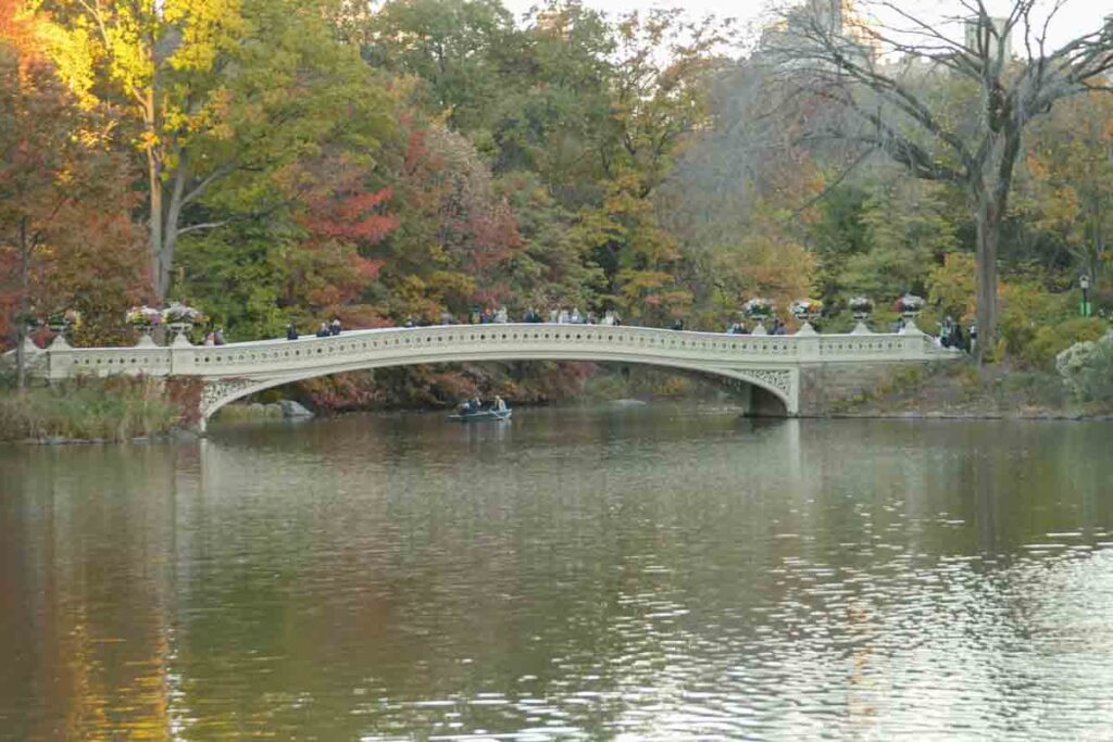 A serene scene of a gently arched bridge with intricate railings spans over a calm body of water. The surrounding trees display autumn colors of orange, red, and yellow. Several people walk on the bridge, and a small boat floats beneath it—an ideal place to visit in the fall.