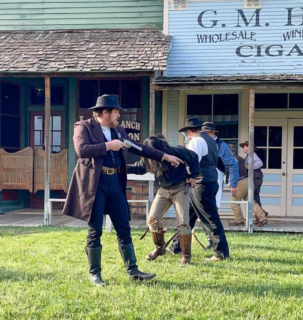 Four people in cowboy attire reenact a Wild West scene outside wooden storefronts; one man points a gun while another hunches forward as if in pain—classic things to do in Dodge City, KS, where the Old West comes to life.