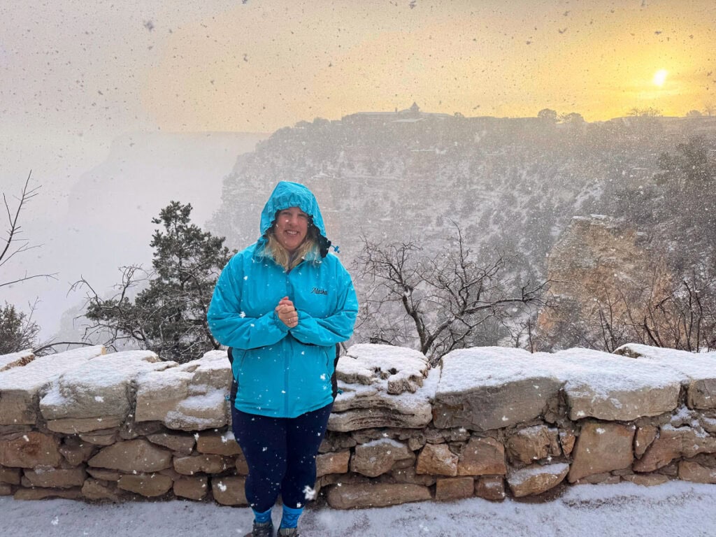 A person in a bright blue jacket stands smiling in a snowy landscape, with a stone wall before them. Behind, one of the best national parks for teens reveals its canyon and trees as a soft sunset casts a warm glow through the gentle snowfall.