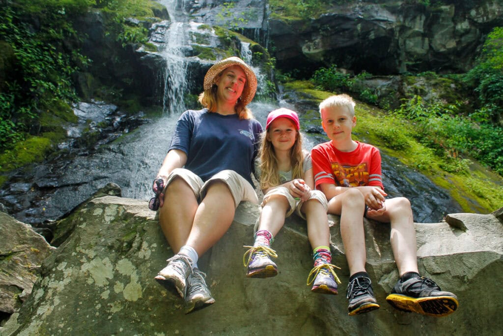 A woman and two children sit on a large rock in front of a majestic waterfall, one of the best national parks for teens to explore. The woman dons a hat and dark shirt, while the children sport red hats and shirts amidst lush greenery and moss-covered rocks.