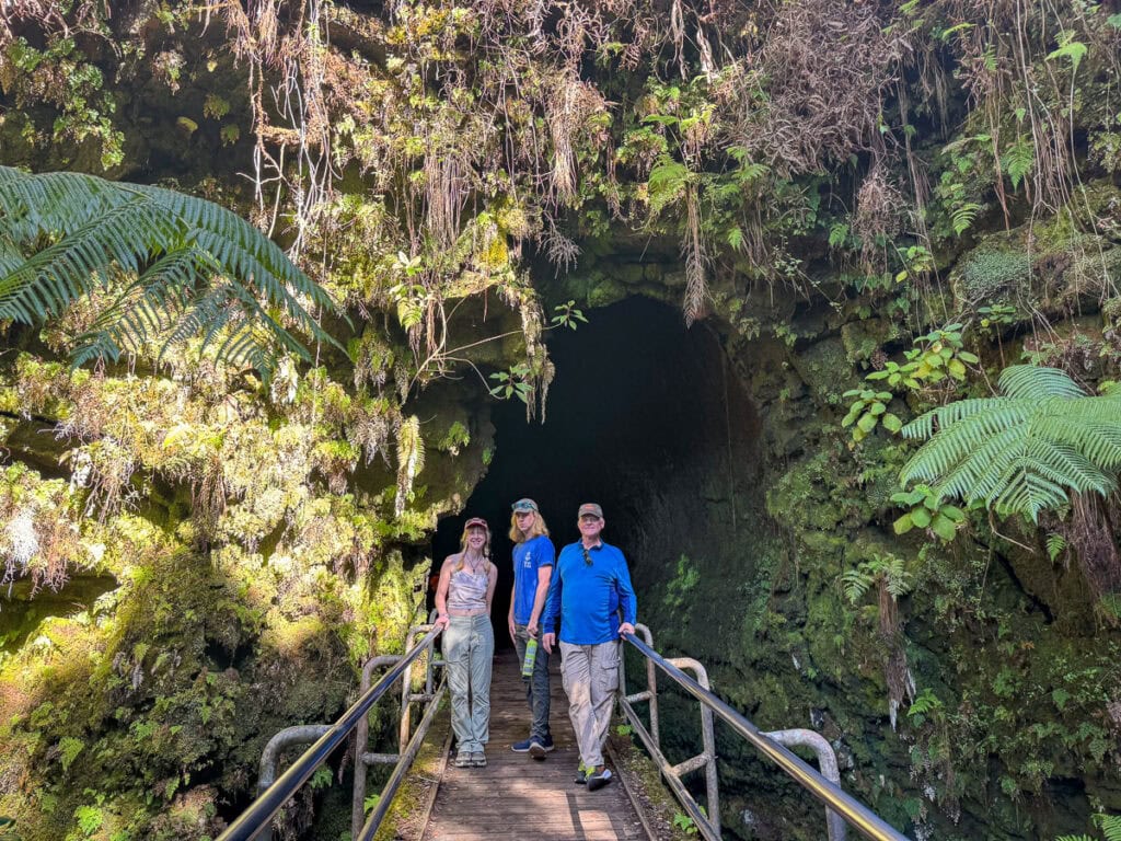 Three people stand on a wooden bridge at the entrance of a mossy, fern-covered cave in one of the best national parks for teens. The lush greenery surrounds the dark opening, contrasting vibrant foliage against shadowy depths, suggesting an exciting nature hike awaits them.