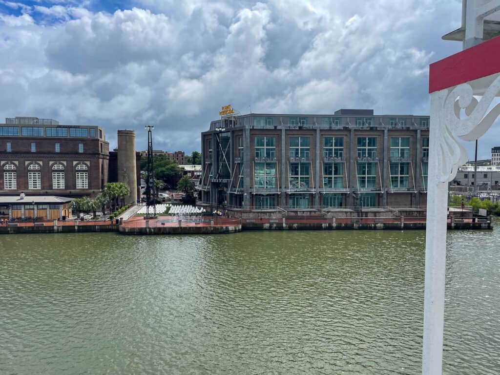 View of a modern glass building and a brick building along a waterfront, with cloudy skies above. Greenish water and a decorative white railing complete this scene near some of the most unique places to stay in Savannah, Georgia.