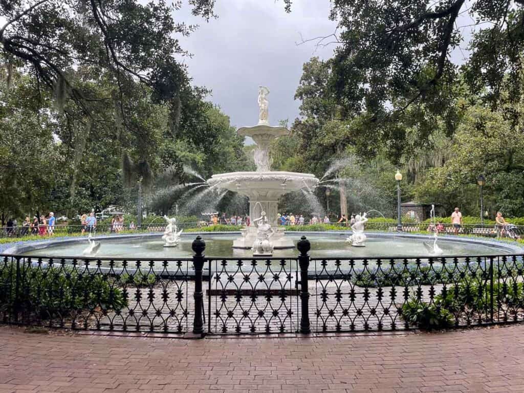 A large white fountain with statues and spraying water stands in the center of a circular pool, surrounded by a black iron fence, trees, and people walking—one of the top things to do in Savannah with teens.
