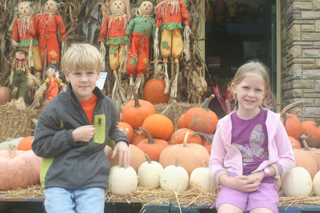 boy and girl in front of a pumpkin patch of multi colored orange pumpkins and decorative scarecrows.