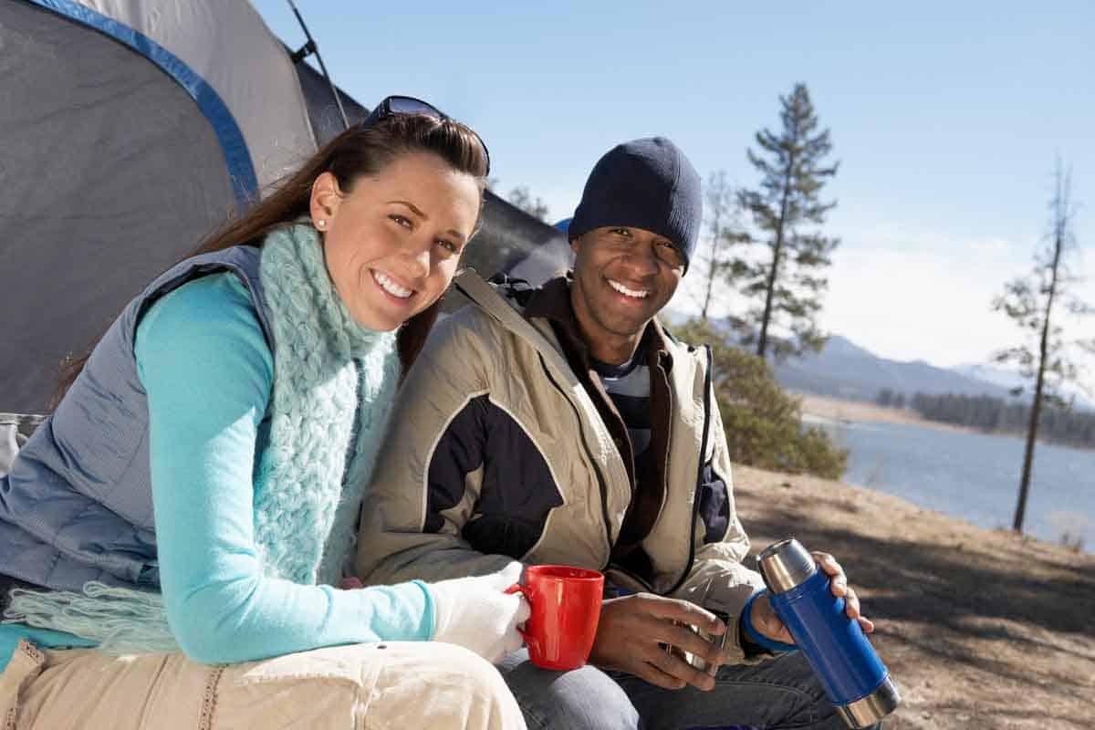 couple sitting in front of a tent with coffee mugs and thermos. lake and mountain in the background with sun shining.