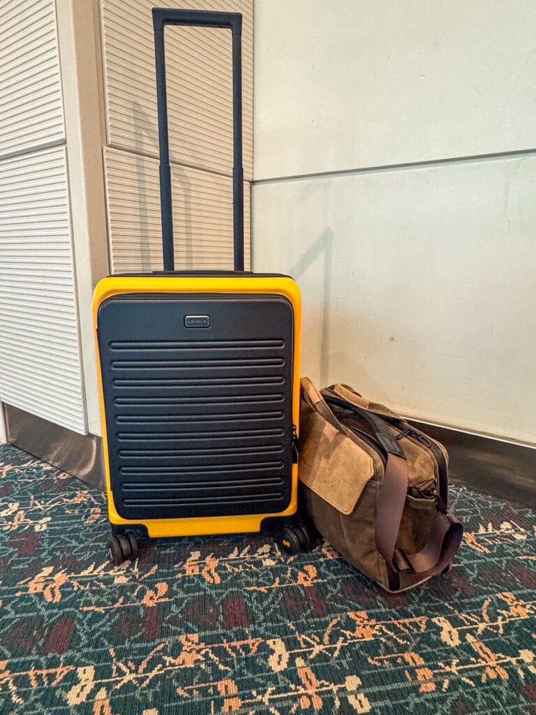 A yellow and black hard-shell suitcase, likely a carry-on vs personal item, stands beside a brown messenger bag on a patterned carpet near a wall, likely in an airport or travel terminal.