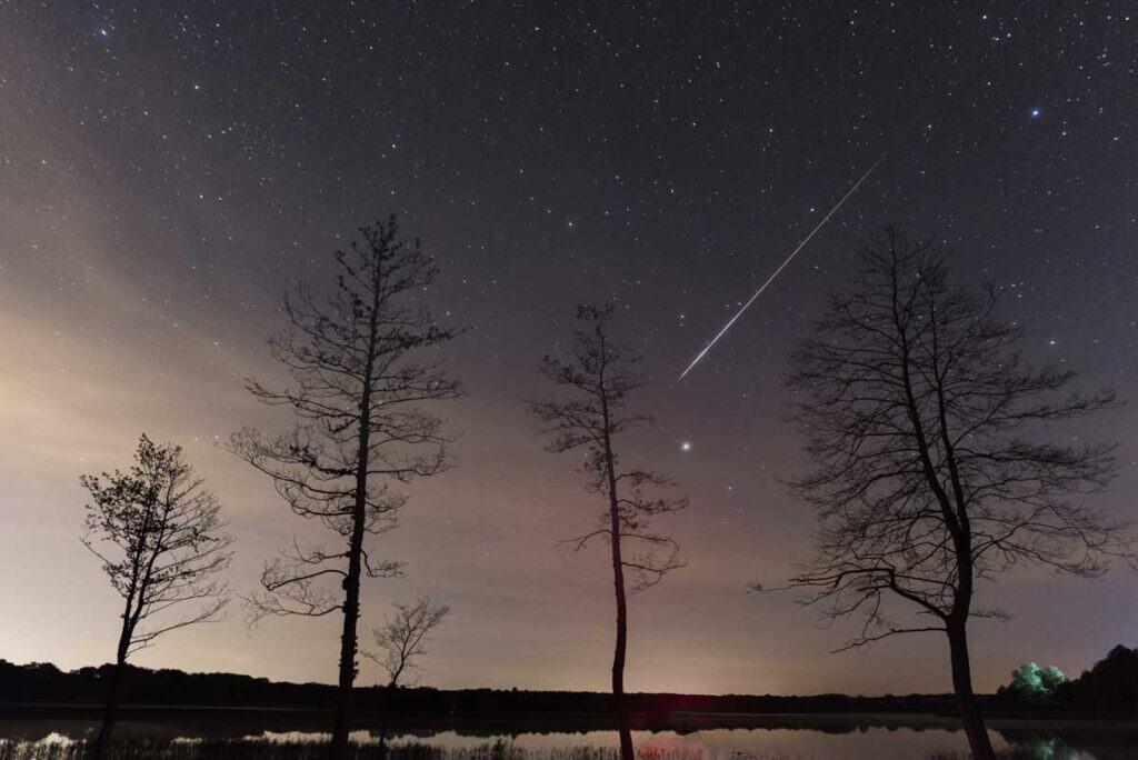 A serene night sky filled with stars over a tranquil lake, with the silhouette of trees in the foreground and a bright meteor streaking through the sky, making it one of the best places to watch a meteor shower