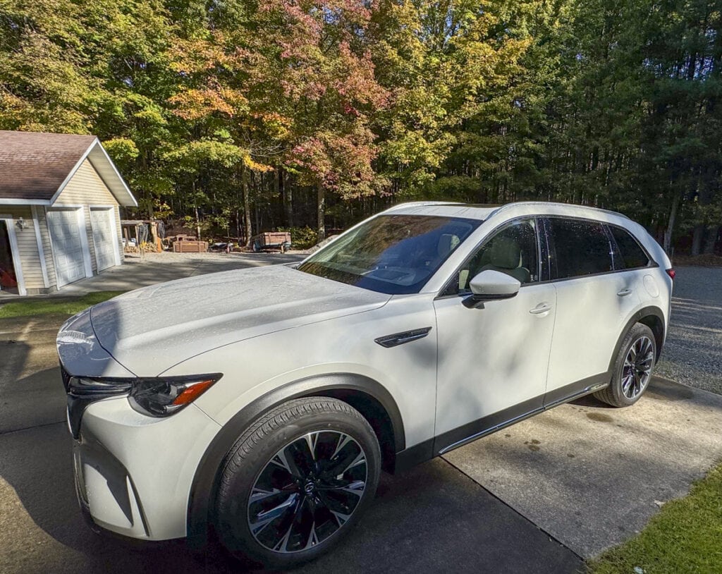 A white Mazda CX-90 SUV is parked on a driveway surrounded by trees, its clean surface and shiny wheels illuminated by the sun. Perfect for a road trip, this scene also features a garage and additional trees in the background.