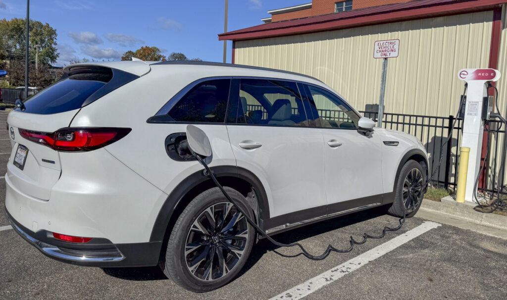 A sleek white Mazda CX-90 is parked at an electric vehicle charging station, plugged in and charging. It occupies a designated space marked "Electric Vehicles Only," perfect for a road trip recharge. In the backdrop, a beige and red building adds to the scene’s contemporary vibe.