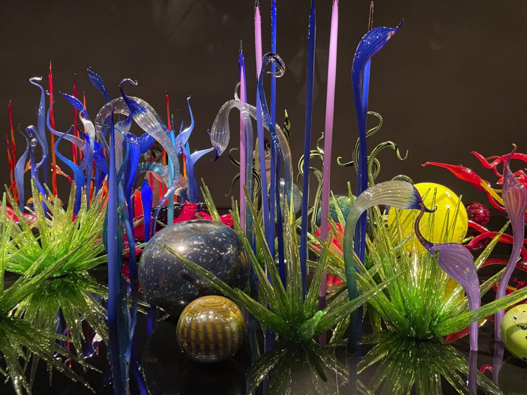 The colorful glass art installation is truly worth it, featuring tall, slender blue and purple glass structures with swirling designs. Green spiky forms spread across the base, alongside polished spheres with intricate patterns, all set against a dark background for a mesmerizing display.