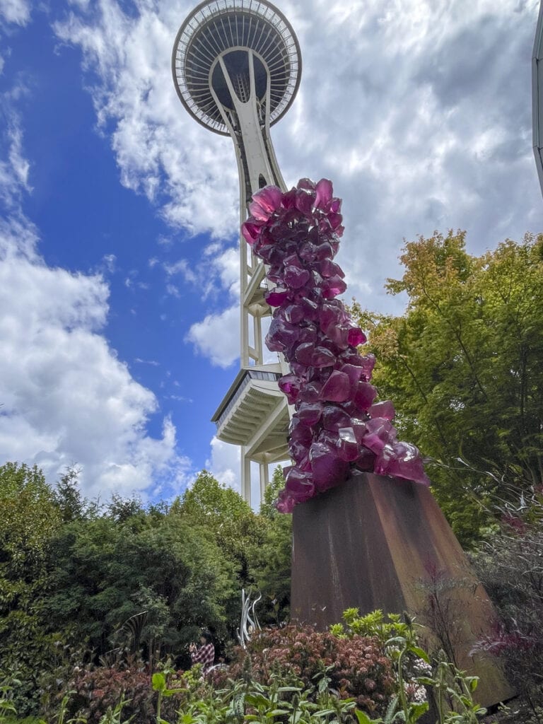 The image captures the iconic Space Needle in Seattle, viewed from below. In the foreground, a tall sculpture of vibrant purple flowers on a metal pedestal stands surrounded by lush greenery under a partly cloudy sky—truly a sight that’s worth it for any CityPASS holder.