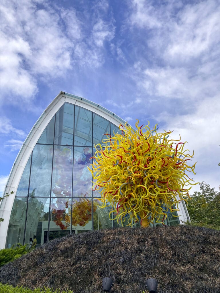 A vibrant outdoor sculpture featuring swirling yellow and orange glass strands stands in front of a modern glass building with a curved roof. The partly cloudy sky enhances the vivid colors, making it a sight worth exploring—and perhaps even included in your CityPASS itinerary.