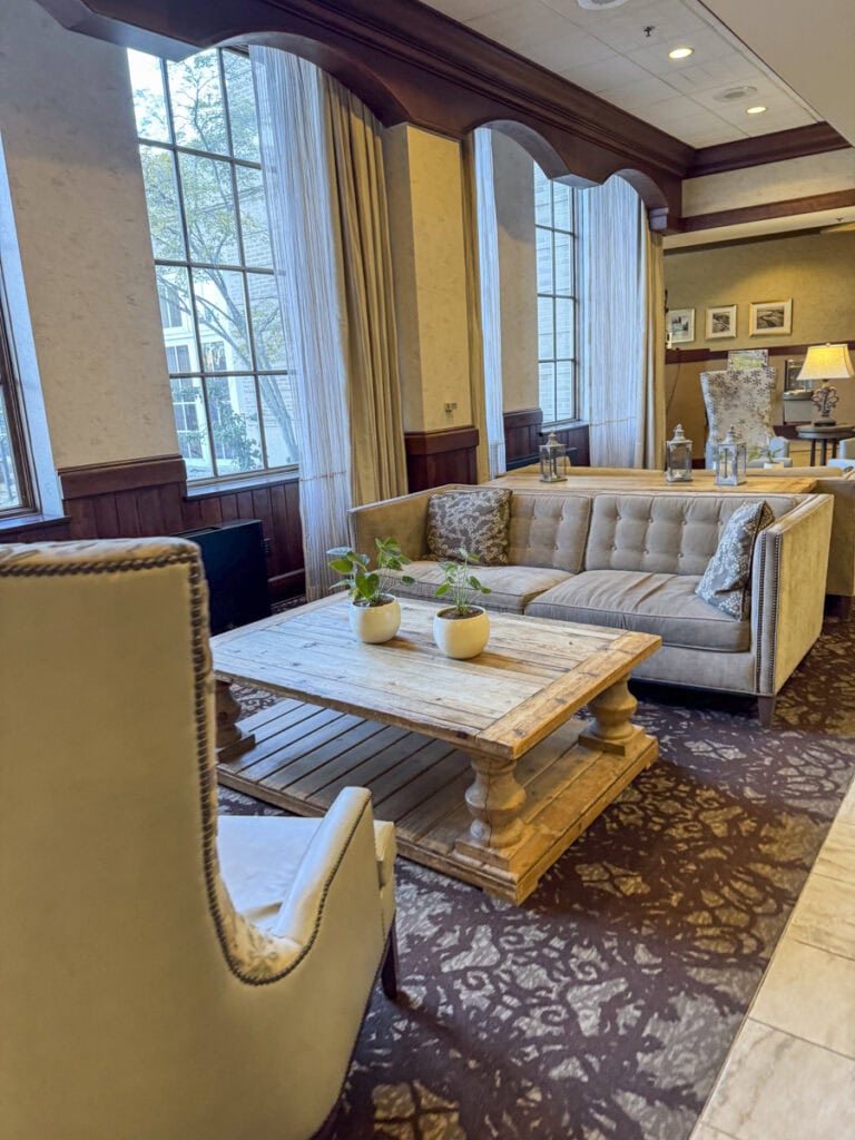 A cozy sitting area at the Park Place Hotel Traverse City features a beige tufted sofa, an armchair, and a wooden coffee table with potted plants. Large windows allow natural light to fill the space, decorated with patterned carpet and elegant curtains for a charming ambiance.