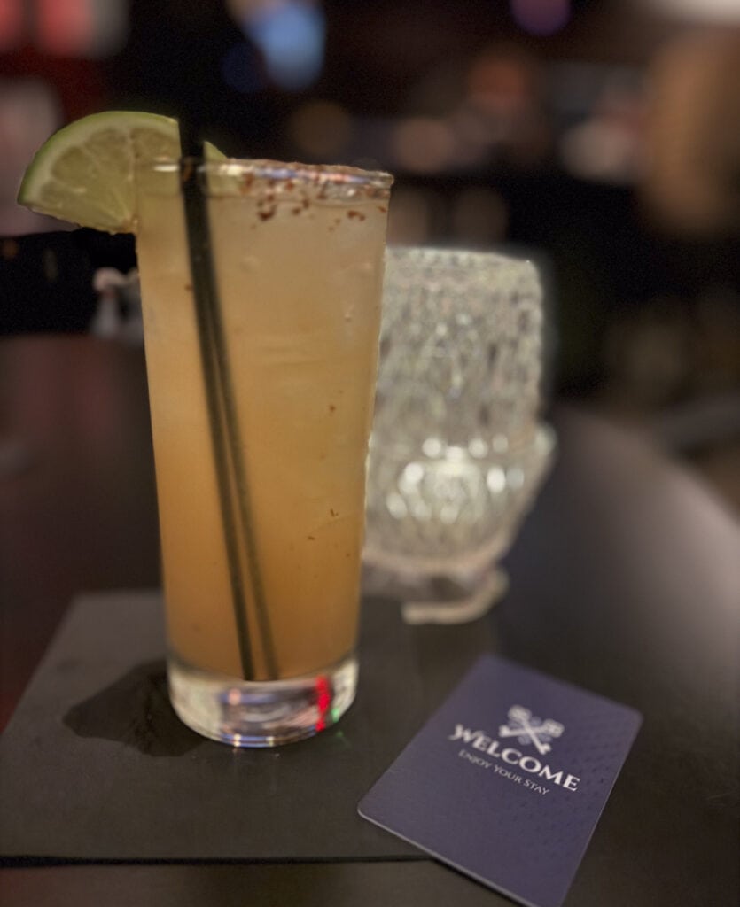A tall glass with a light brown cocktail, garnished with a lime slice and two black straws, sits on a dark table at Park Place Hotel Traverse City. A textured glass and a “Welcome” card are blurred in the background, suggesting a cozy restaurant or bar setting.