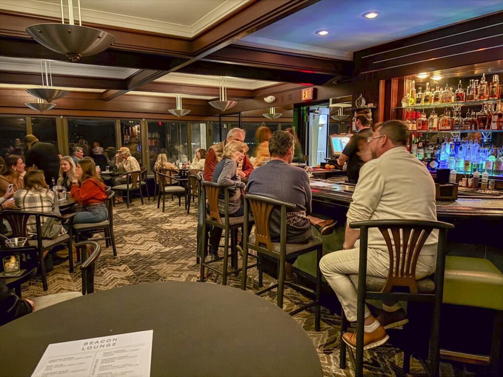 Nestled in the heart of the Park Place Hotel Traverse City, this cozy bar invites patrons to unwind on bar stools or at tables, engaged in lively conversation. The shelves behind the bar brim with various bottles, and warm, inviting lighting enhances the relaxed atmosphere.