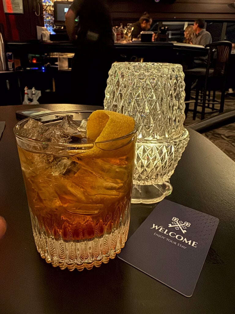 A glass of whiskey on the rocks with a lemon twist rests on a dark table at Park Place Hotel Traverse City, accompanied by a textured glass light fixture and a "Welcome" card. The dimly lit bar setting adds to the elegant ambiance.