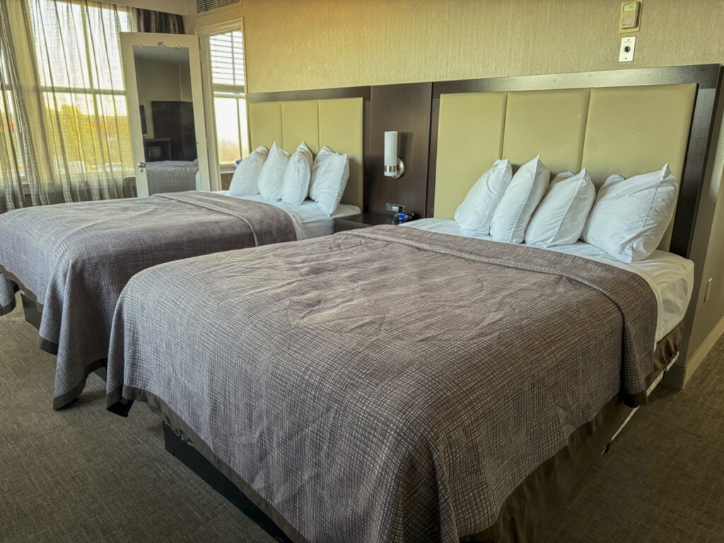 At Park Place Hotel in Traverse City, the room features two neatly made double beds adorned with gray-brown blankets and four plush pillows each. Natural light streams through sheer curtains, while a small TV in the corner complements the room's modern decor.