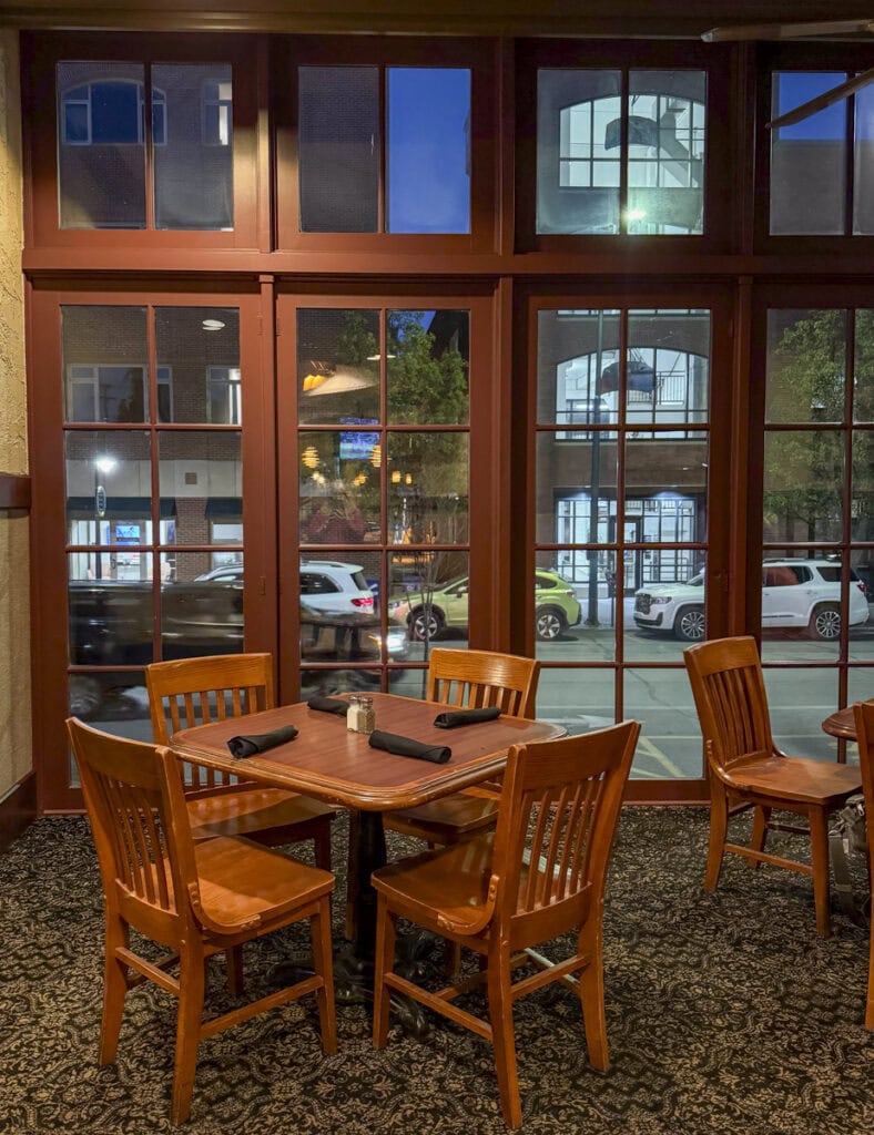 A cozy restaurant at Park Place Hotel in Traverse City showcases a wooden table set for four with dark napkins by large windows. Outside, the parking lot and city buildings are beautifully illuminated by the evening sky.