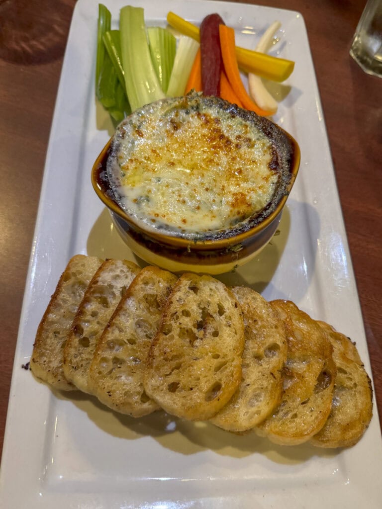 A dish with baked cheese dip in a bowl, garnished with herbs, served on a white rectangular plate with slices of toasted bread and fresh vegetable sticks, including celery and carrots. Perfect for pairing with the elegant ambiance at Park Place Hotel Traverse City.