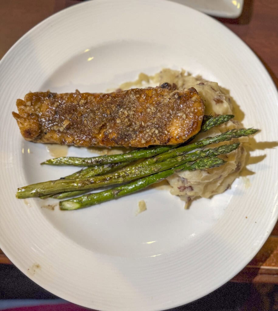 A plate from Park Place Hotel Traverse City showcases a grilled salmon fillet topped with seasoning, served alongside three asparagus spears and a mound of mashed potatoes. The exquisite meal is elegantly presented on a white ceramic plate.