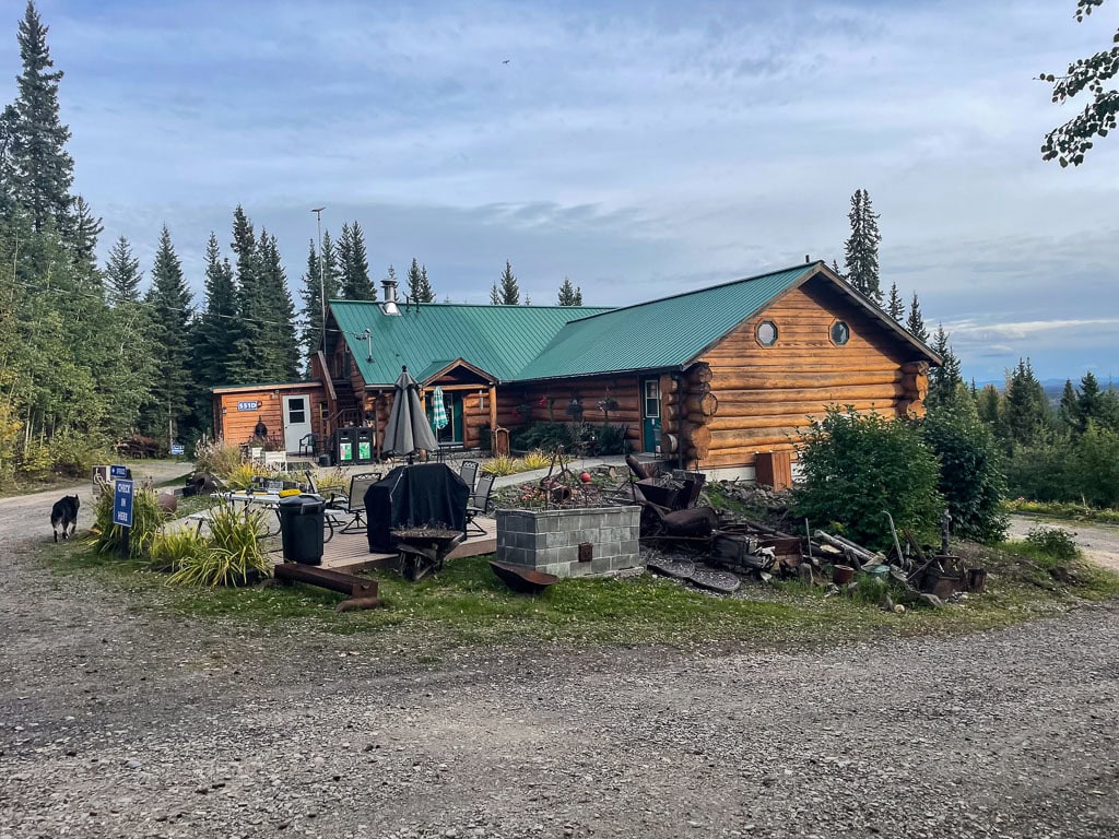 Nestled amidst pine trees, this log cabin with a green metal roof offers a serene retreat. A gravel driveway meanders to a fire pit with chairs, perfect for evenings under overcast skies. Unlike hotels in Fairbanks, Alaska, enjoy the rustic charm while your dog explores the surroundings.