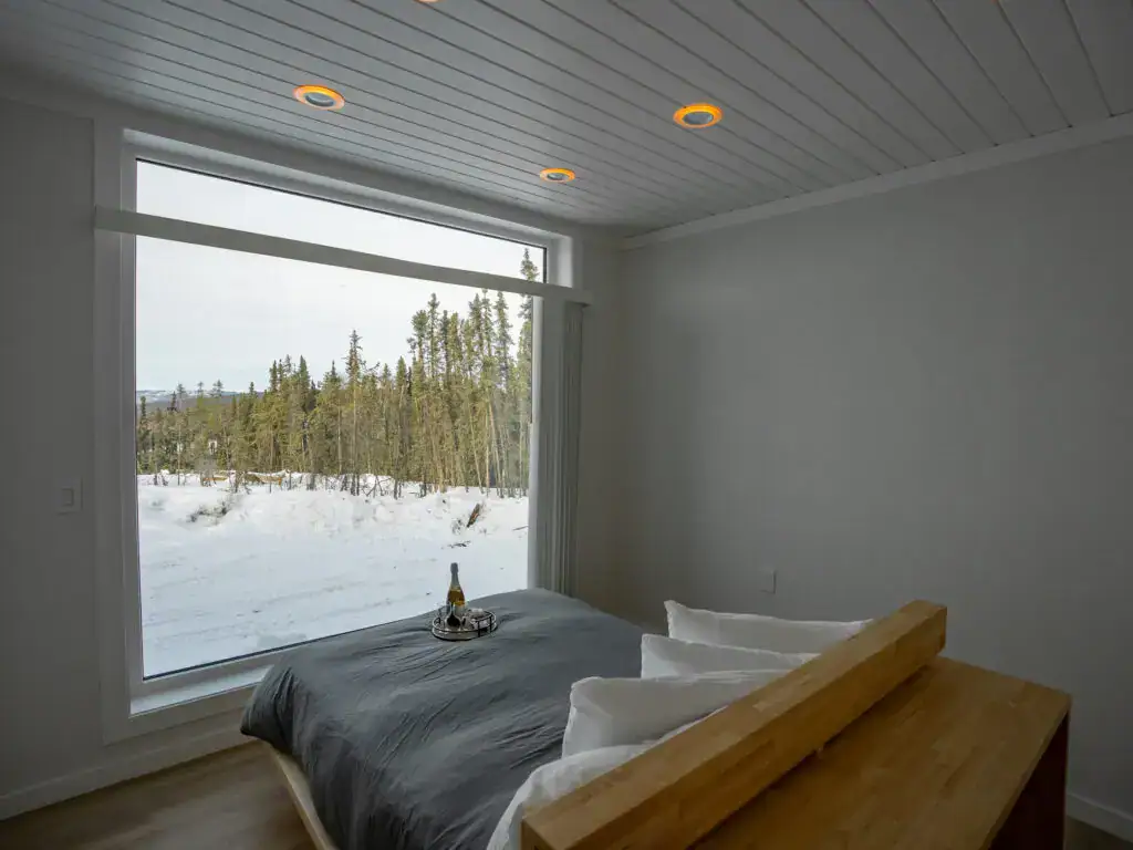 A minimalist bedroom in the style of hotels in Fairbanks, Alaska offers a vast snowy forest view through a large window. A wooden bed with grey bedding faces this serene scene. Nearby, a tray with a bottle and glass rests on the bed, highlighted by recessed ceiling lights on pristine white walls.