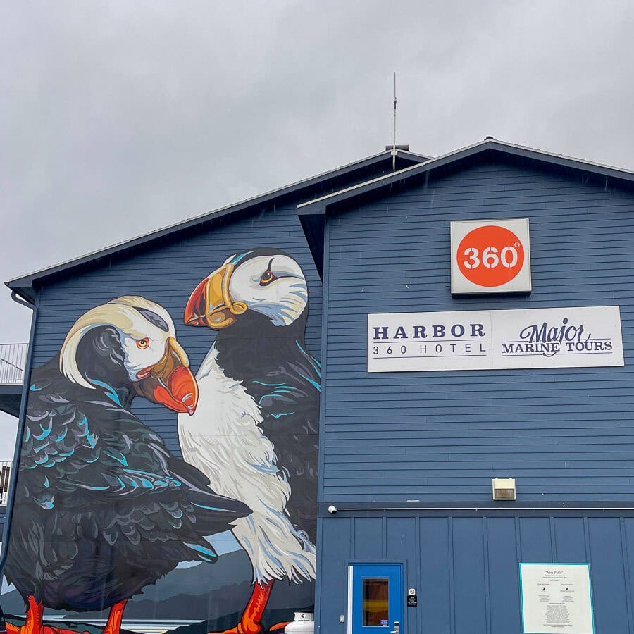 The image shows a large mural of two puffins on the side of a blue building, inviting you to plan a trip to Alaska. Signs for "Harbor 360 Hotel" and "Major Marine Tours" beckon travelers, while a circular orange "360ยฐ" logo hangs above. The sky is gray and overcast.