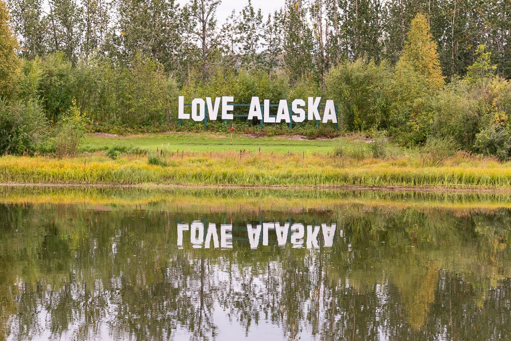 A scenic view featuring the words "LOVE ALASKA" displayed in large white letters amidst lush greenery. The text is reflected on the calm surface of a pond in the foreground, with tall trees in the background. The Love Alaska Sign is at Pike's Waterfront Lodge, one of the best hotels in Fairbanks to see Northern Lights
