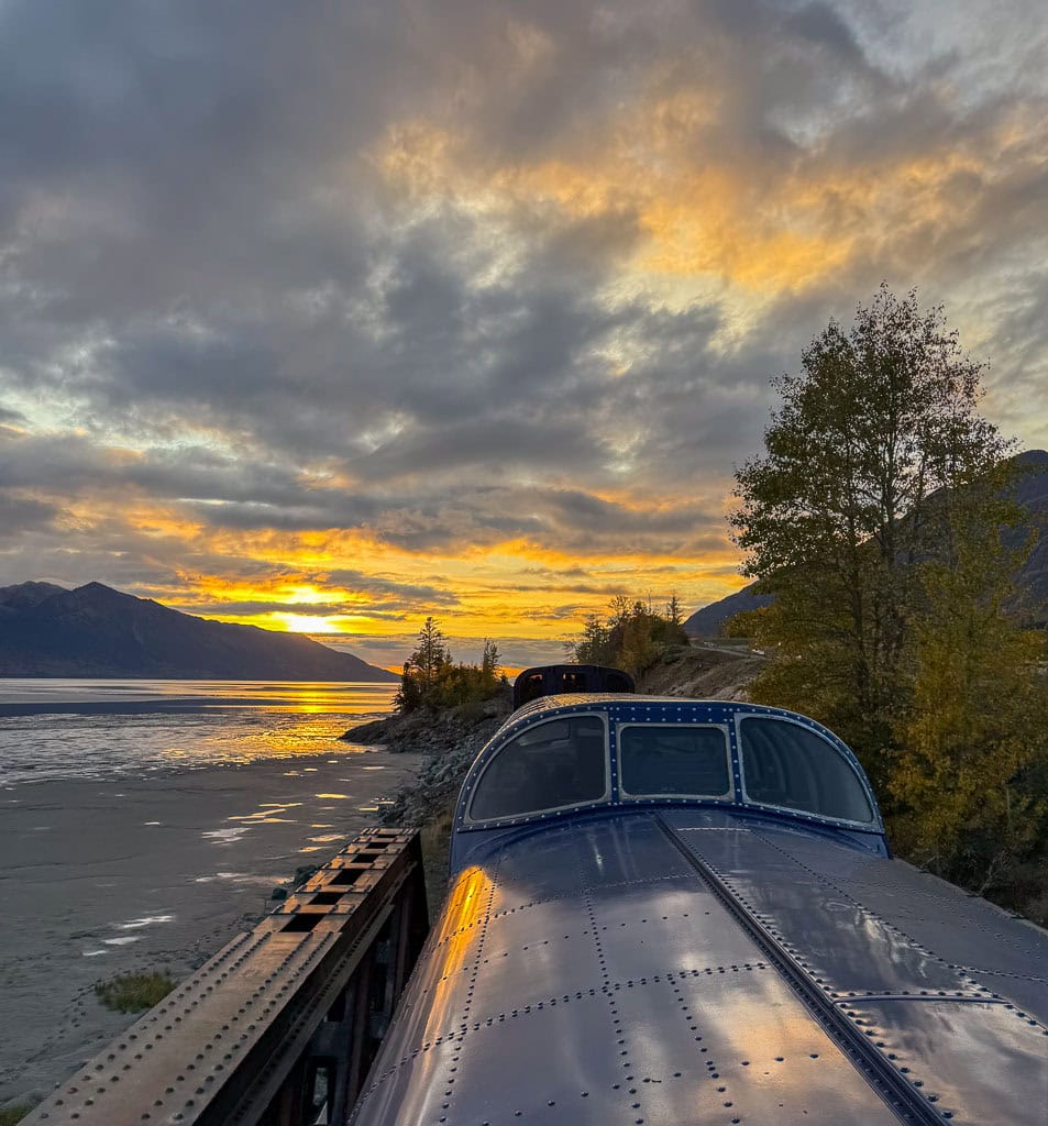 A train travels along a scenic route during sunset, reminiscent of planning a trip to Alaska. The sun illuminates the sky with golden hues, reflecting on the water below. Mountains and trees border the body of water, creating a picturesque autumn landscape.