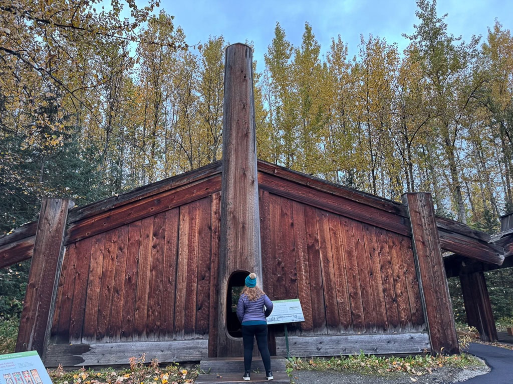 A person wearing a blue jacket and a green beanie stands in front of a large wooden structure with a triangular roof, reminiscent of those you'd find when you plan a trip to Alaska. The backdrop features tall trees with autumn leaves, and a sign is placed in front of the structure.
