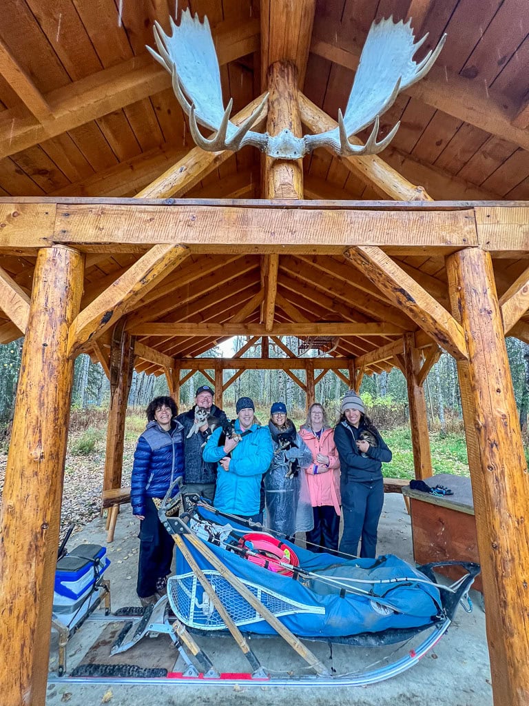 A group of people stands in a wooden pavilion adorned with moose antlers, dressed for winter. A sled loaded with gear sits before them, while two hold dogs. The backdrop of trees and grass whispers the promise of adventureโperhaps a call to plan a trip to Alaska.