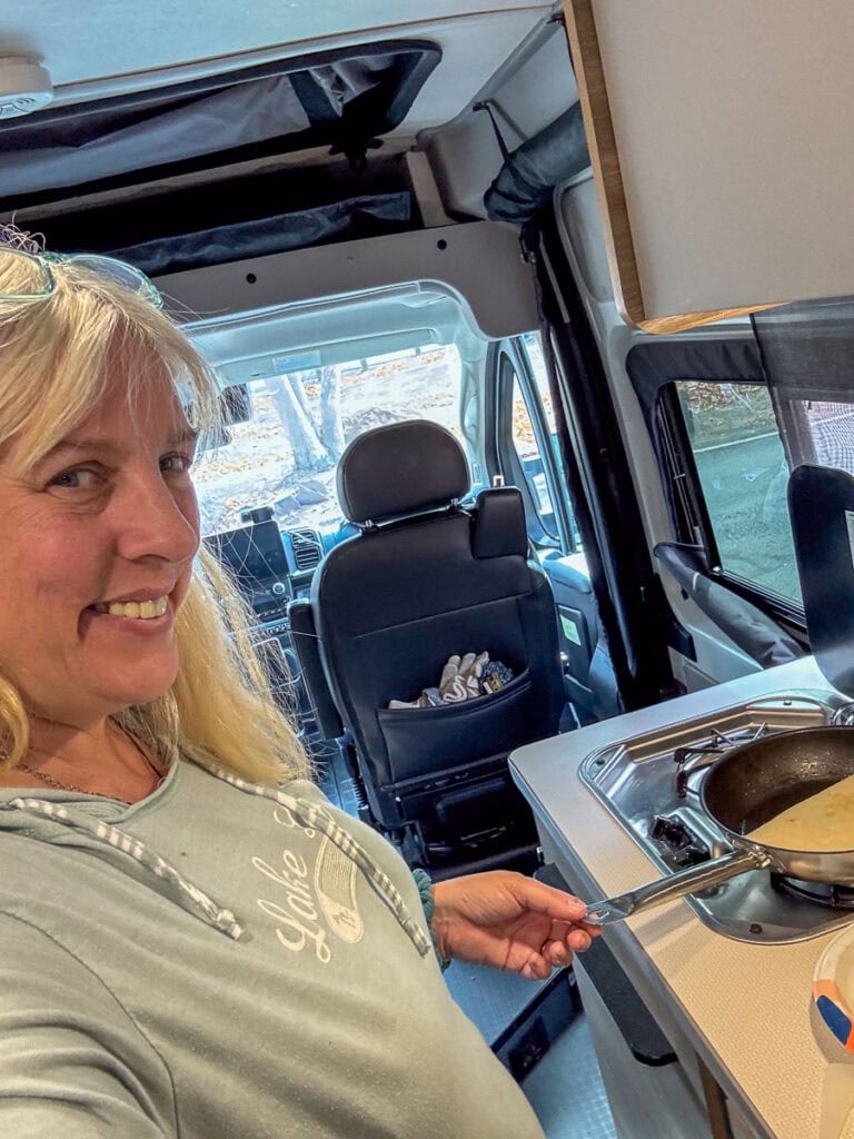A person with blonde hair is smiling while cooking an omelet in a frying pan inside a camper van. The bright and spacious interior hints at a comfortable adventure, perfect for anyone considering Blacksford RV Rental for their next road trip. They're wearing a light blue sweatshirt, adding to the cozy vibe.