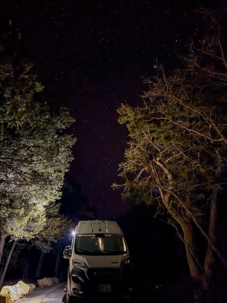 A white camper van, from a glowing Blacksford RV Rental review, is parked on a dark wooded road under a starry night sky. Trees frame the van as their branches reach upward, with soft light illuminating the scene, highlighting the serene night setting.