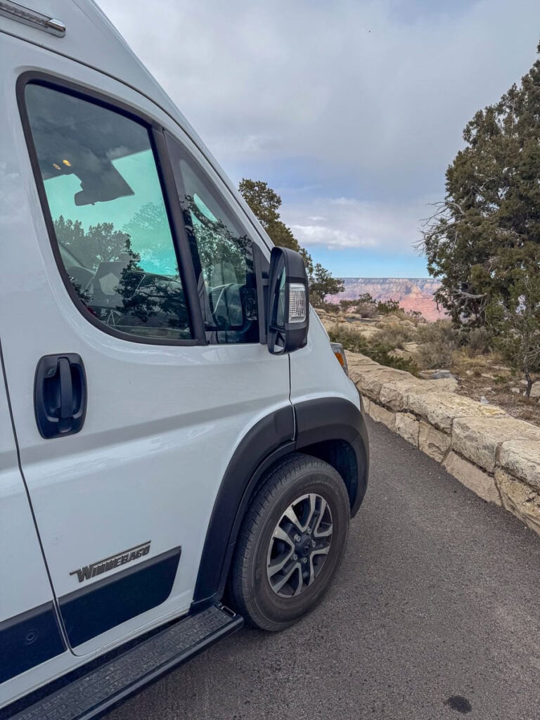 A sleek white camper van from Blacksford RV Rental is parked on a paved road beside a stone barrier, offering an idyllic backdrop of a scenic canyon and lush trees beneath a partly cloudy sky.