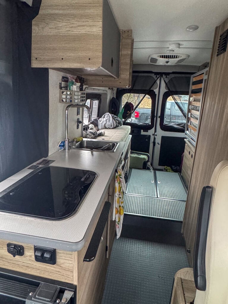 The interior of the camper van from Blacksford RV Rental showcases a compact kitchen with a sink, stove, and wooden cabinets. A gray countertop leads to a cozy seating area at the rear. Through partially visible windows, an exterior view hints at adventure awaiting beyond.