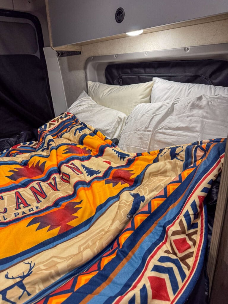 Cozy bed setup inside a camper van, featuring white pillows and a colorful, patterned blanket with "Canyon" text. An overhead light casts a warm glow, highlighting the compact and inviting sleeping area. Ideal for those inspired by glowing Blacksford RV Rental reviews.