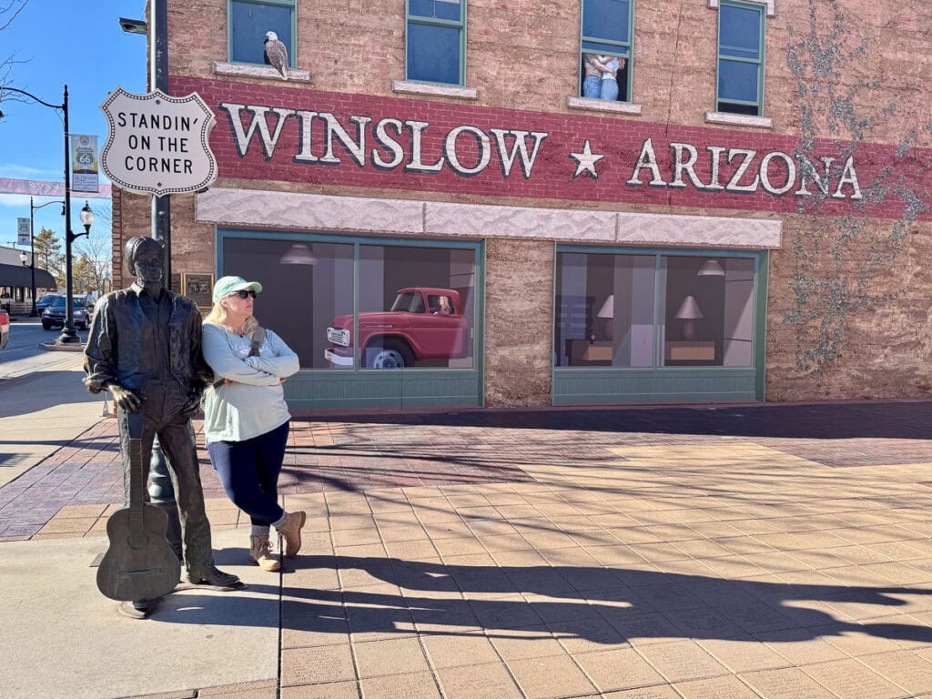 A woman poses with a statue of a man holding a guitar in front of a mural and the "Winslow Arizona" sign, capturing the spirit of travel akin to embarking on an adventure after reading glowing Blacksford RV Rental reviews. A red truck and two figures peek from the mural's windows, adding charm to the scene.