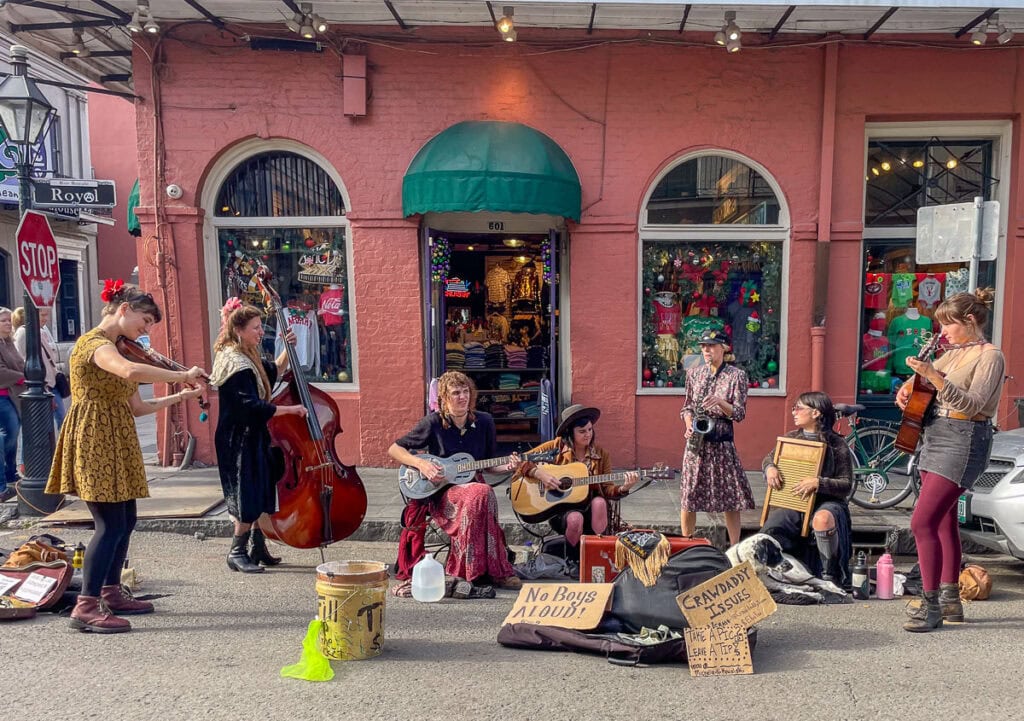 A group of six street musicians, mostly women, perform in front of a pink building—one of the vibrant things to do in New Orleans with teens. They play violin, upright bass, and more, with tip jars and open instrument cases set before them.