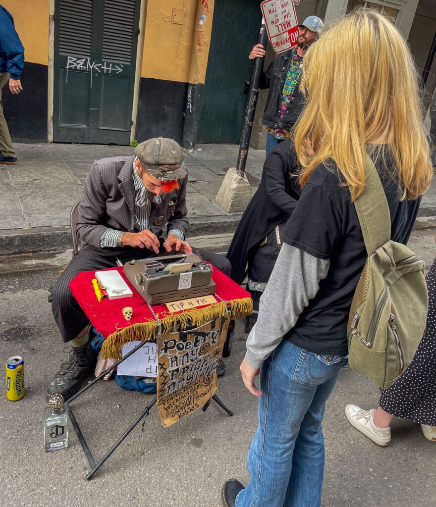 A street poet in a pinstripe suit and hat types on a vintage typewriter at a small table, while a woman with blonde hair and a green backpack watches—one of the unique things to do in New Orleans with teens on a lively city sidewalk.