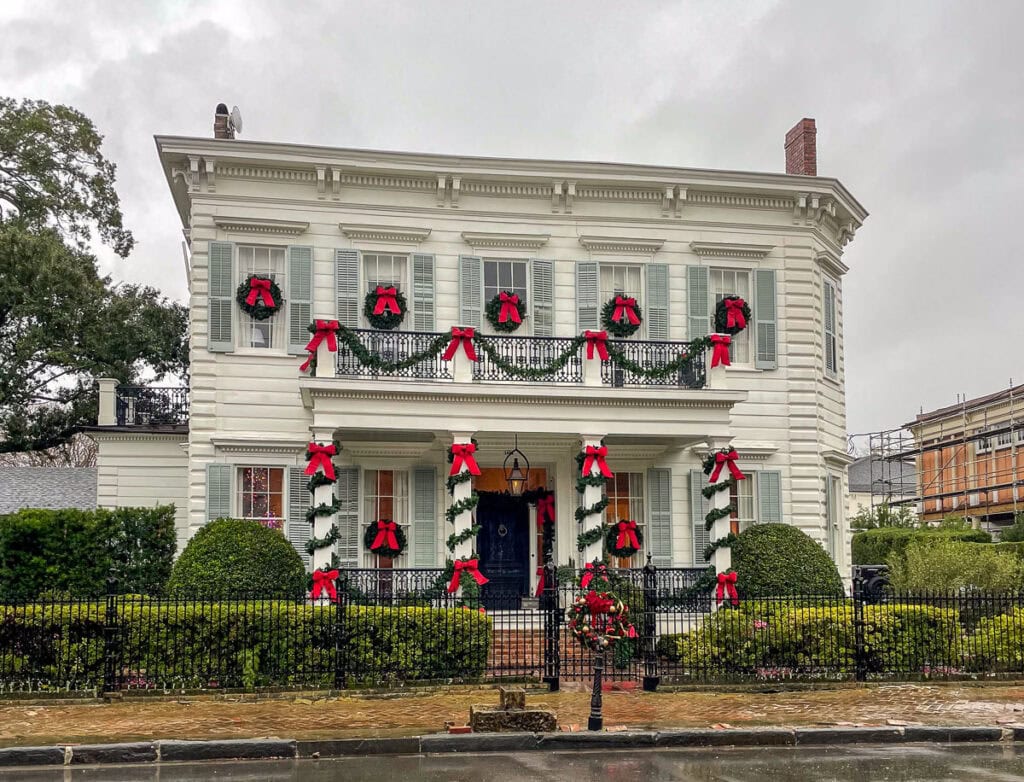 A two-story white house decorated for the holidays with red bows, green garlands, and wreaths on the balcony and windows—a festive scene often admired on rainy days behind a black iron fence. A charming sight among things to do in New Orleans with teens.