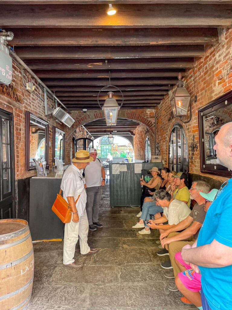 A group of people sit on a bench inside a rustic, brick-walled corridor with wooden beams—one of many things to do in New Orleans with teens. Two people stand in front, one wearing a hat and orange bag. Large lanterns hang overhead as daylight shines through an archway.